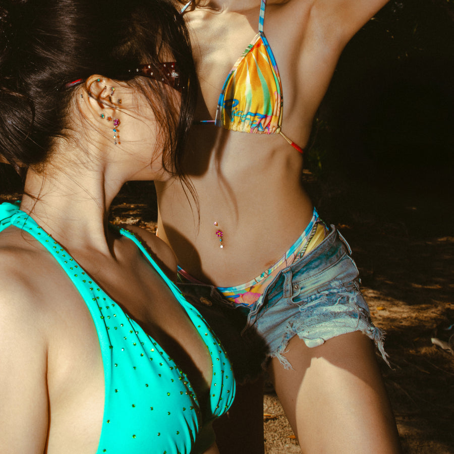 Two people in colorful swimwear standing close together outdoors with colorful ear and belly button jewelry