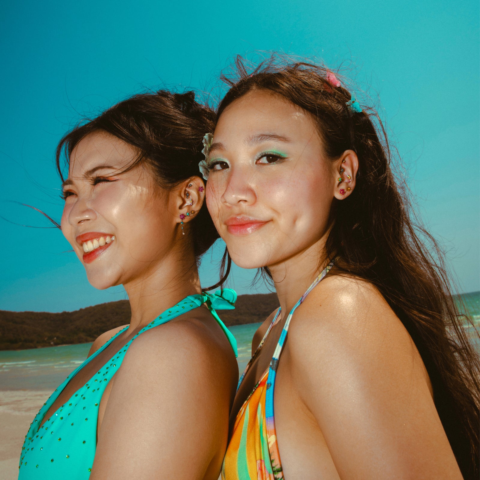 Two women wearing colorful earrings standing on a beach with clear blue sky.
