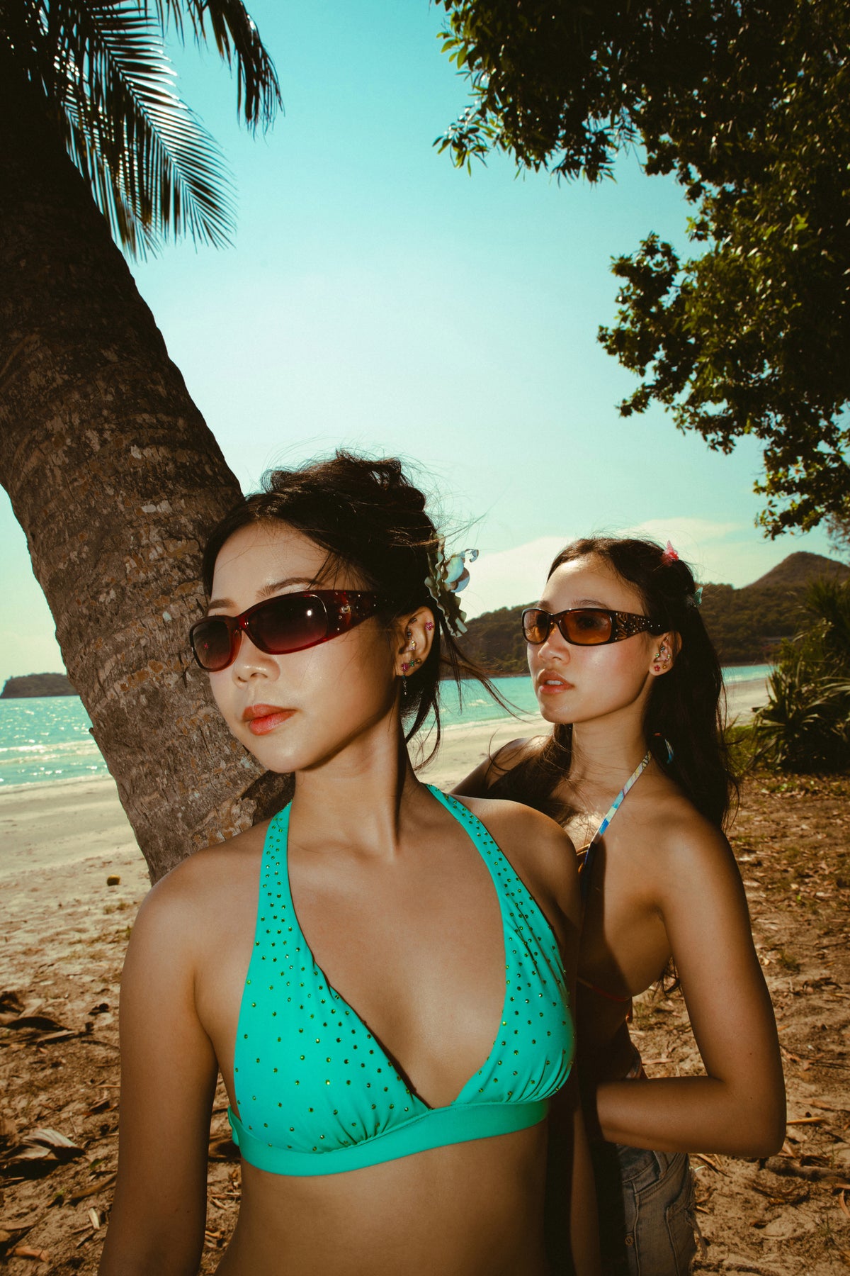 Two models wearing statement earrings and sunglasses on a tropical beach, styled in a Y2K-inspired summer lookbook for Oh Kira!