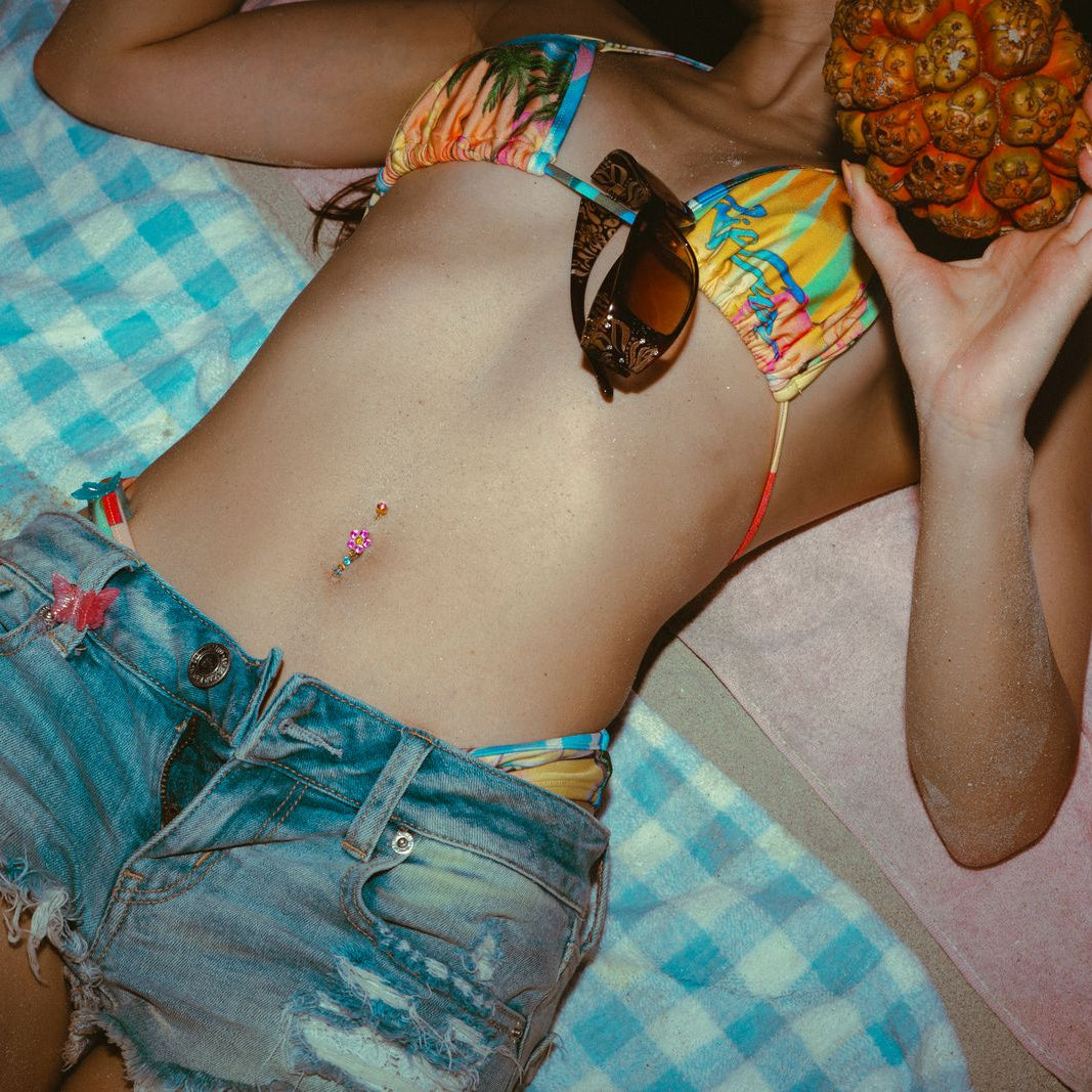 Girl with pink CZ flower belly button jewelry in colorful bikini holding a tropical fruit on a beach blanket