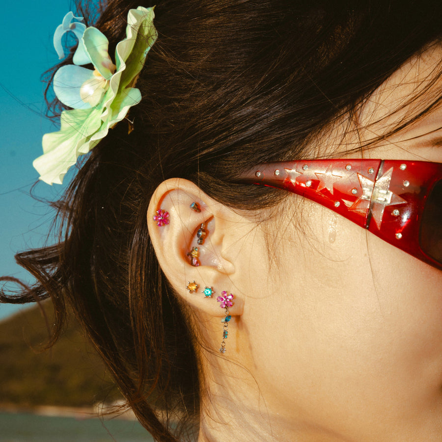 Close-up of an ear with colorful earrings, red sunglasses, and a floral hair accessory against a blue sky.