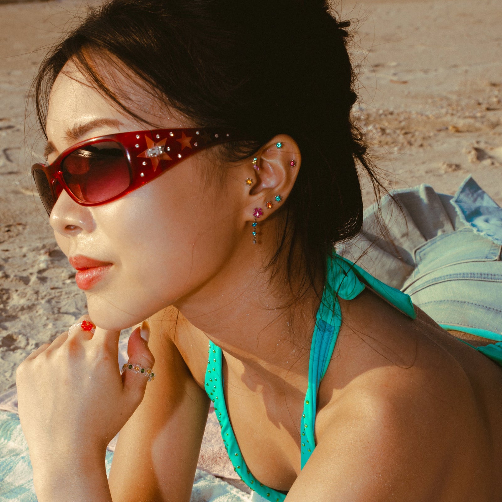 Woman wearing red sunglasses and a turquoise bikini top and colorful earrings on a beach.