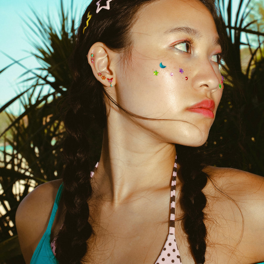Woman with braided hair, colorful earrings, and face gems in front of a tropical background